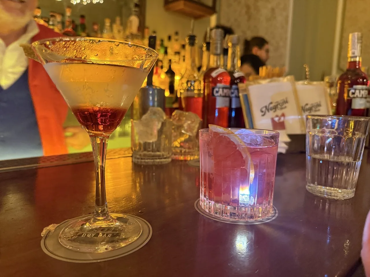 A coupe cocktail and a pink cocktail on a wooden bar counter in front of shelves of bottles