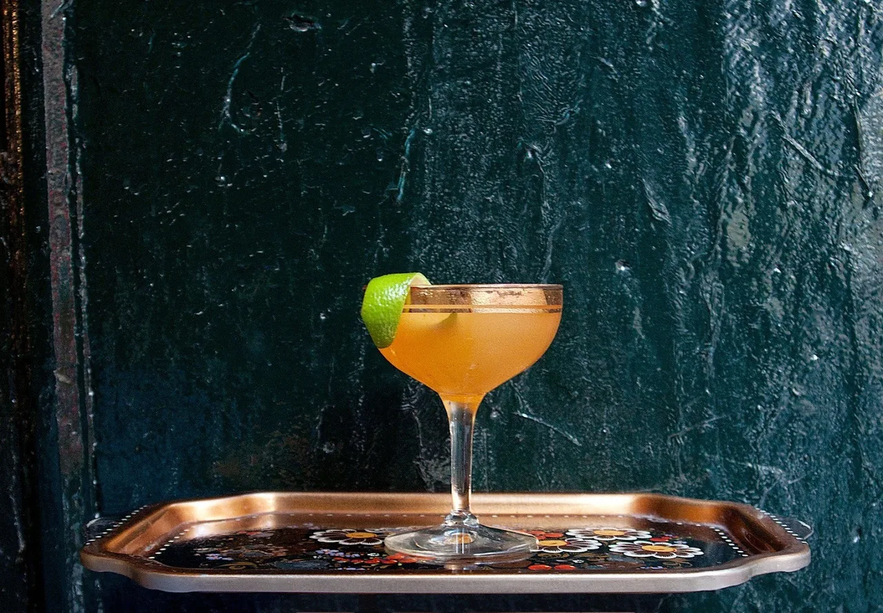 An orange coupe cocktail with a lime wedge on a copper tray against a dark green wall