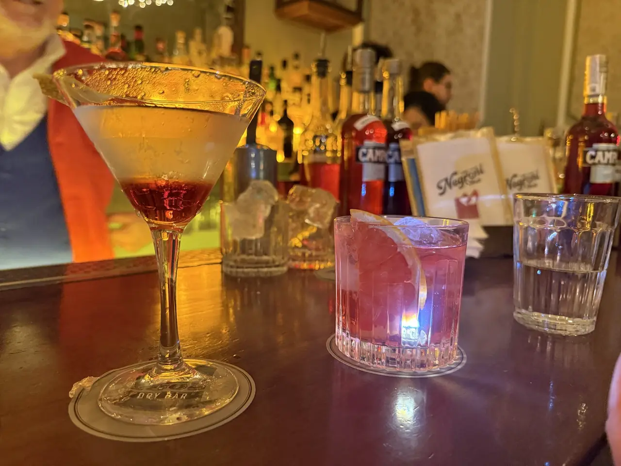 Martini-style cocktail with lemon twist beside a rose-colored cocktail garnished with grapefruit on bar counter
