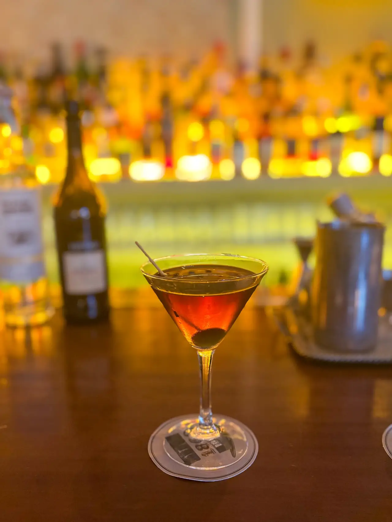 A dark amber cocktail with a cherry garnish in a martini glass on the bar top