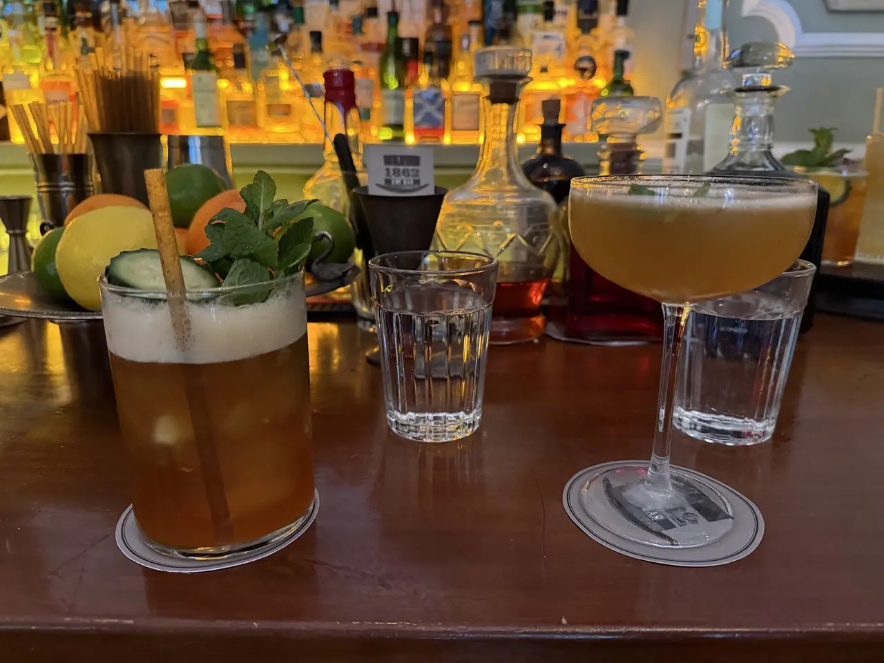 Two cocktails on the bar, one garnished with mint and cucumber, the other a frothy coupe