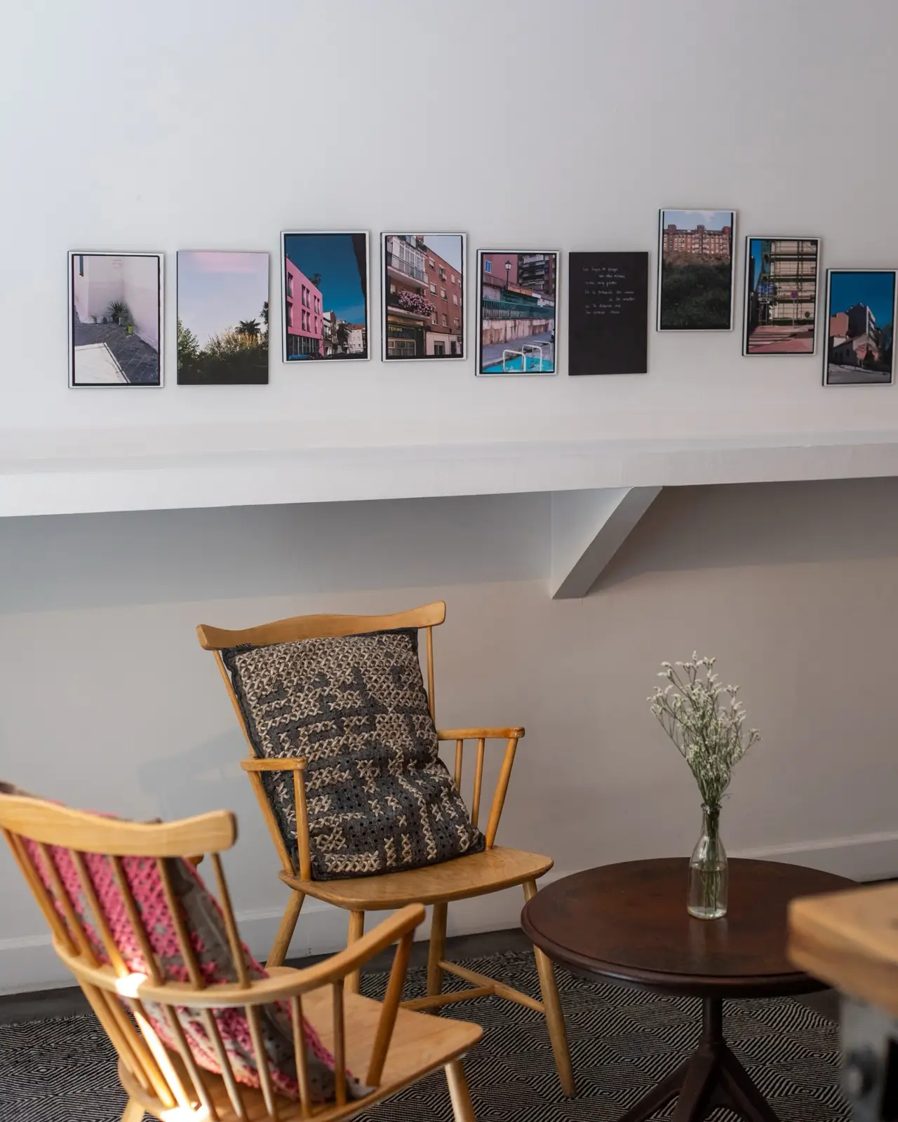 Interior seating corner with wooden spindle-back chairs, patterned cushions, a small round table with a vase, and a row of framed photographs on the wall.