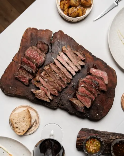 Sliced grilled steak fanned on a charred wooden board with roasted potatoes, grilled bread, and dipping sauces at 9 Reinas