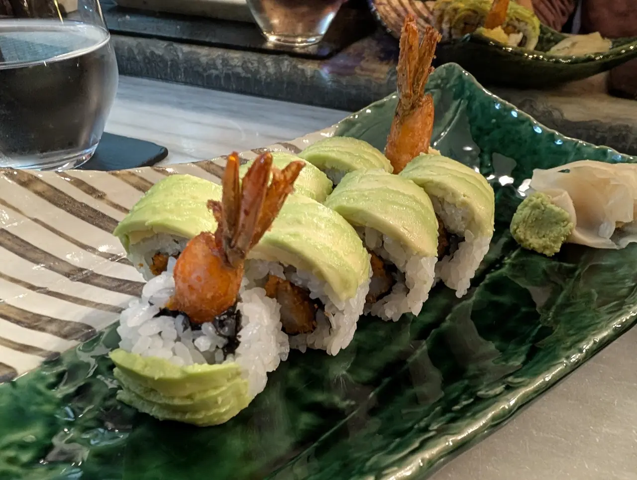 Avocado-topped sushi rolls with tempura shrimp tails standing upright, served with wasabi and pickled ginger