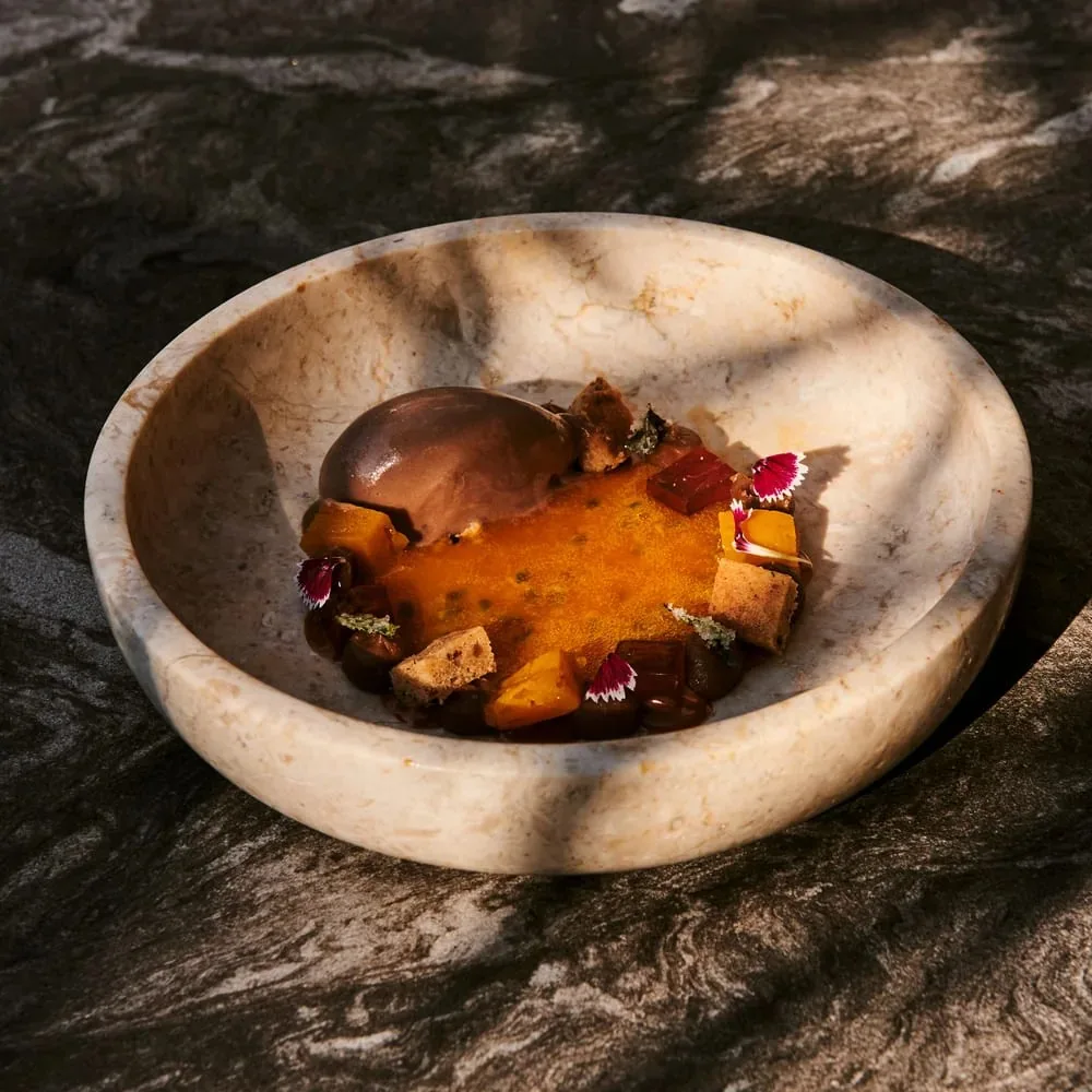 Dessert in a shallow cream marble bowl: a scoop of dark chocolate ice cream beside a smooth orange puree scattered with cubes of jelly, sponge pieces and small