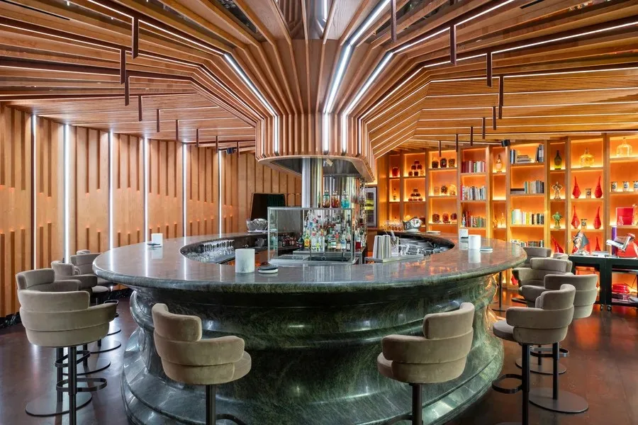Circular green marble bar with velvet stools around it, under a fan of wooden ceiling slats and strip lighting, backed by warmly lit shelves holding bottles, bo