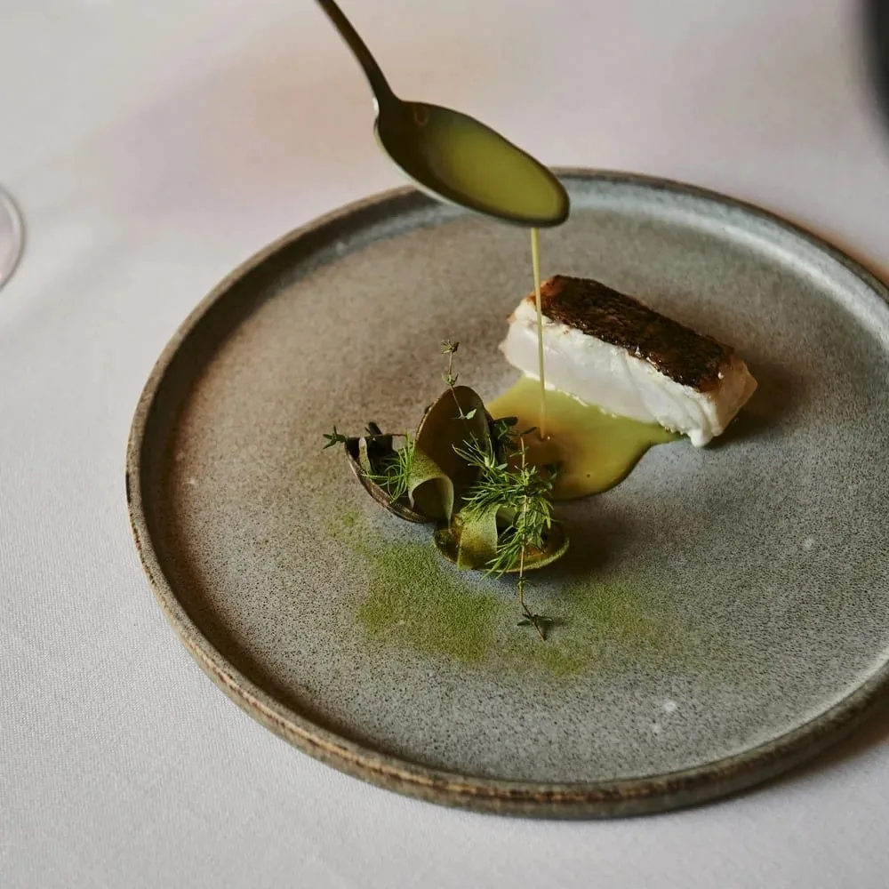 A piece of white fish with a green sauce being poured over it on a grey plate