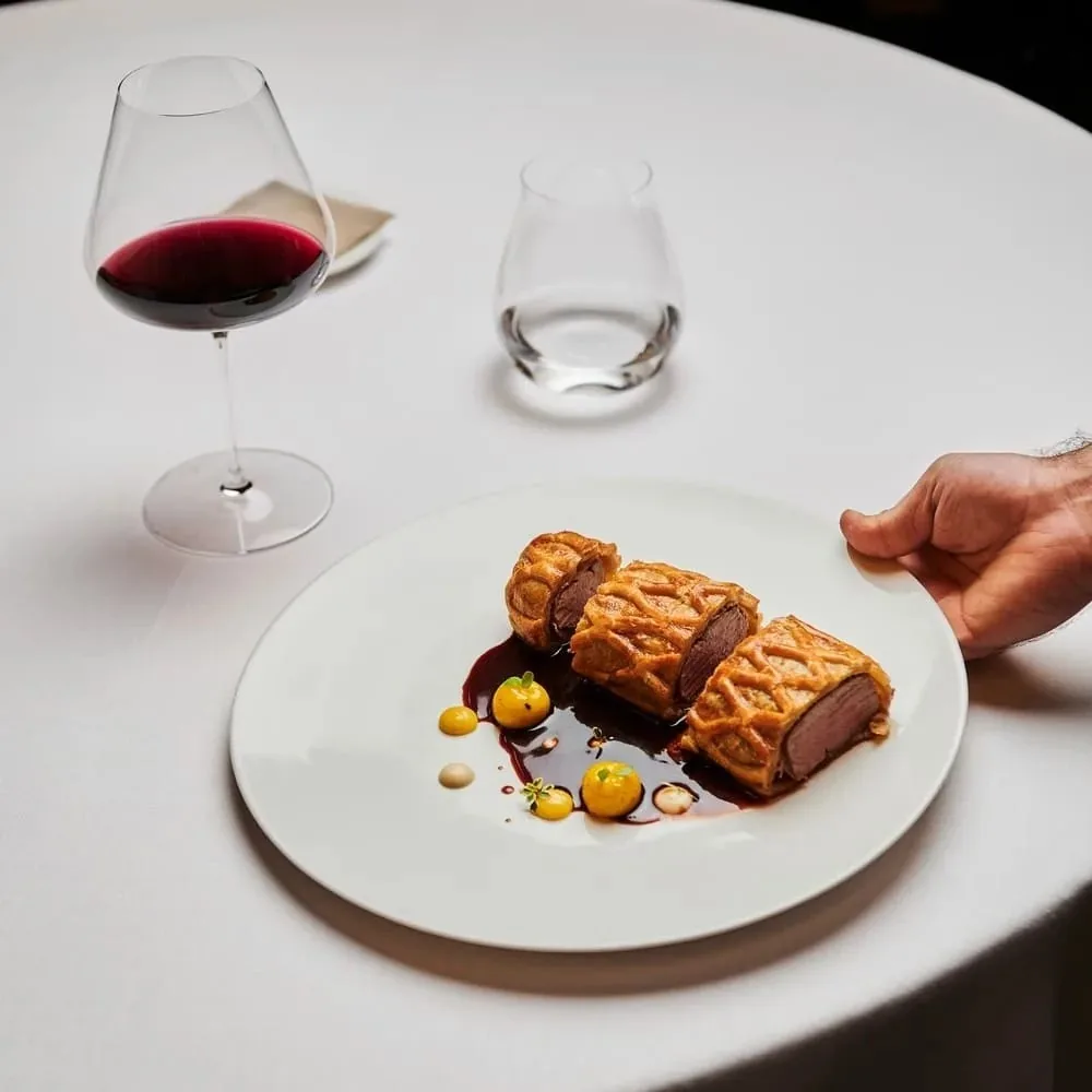 A white plate of two sliced roasted meat rolls with yellow dots of sauce and a glass of red wine