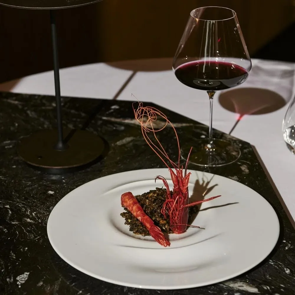 A langoustine over dark grains on a white plate beside a glass of red wine