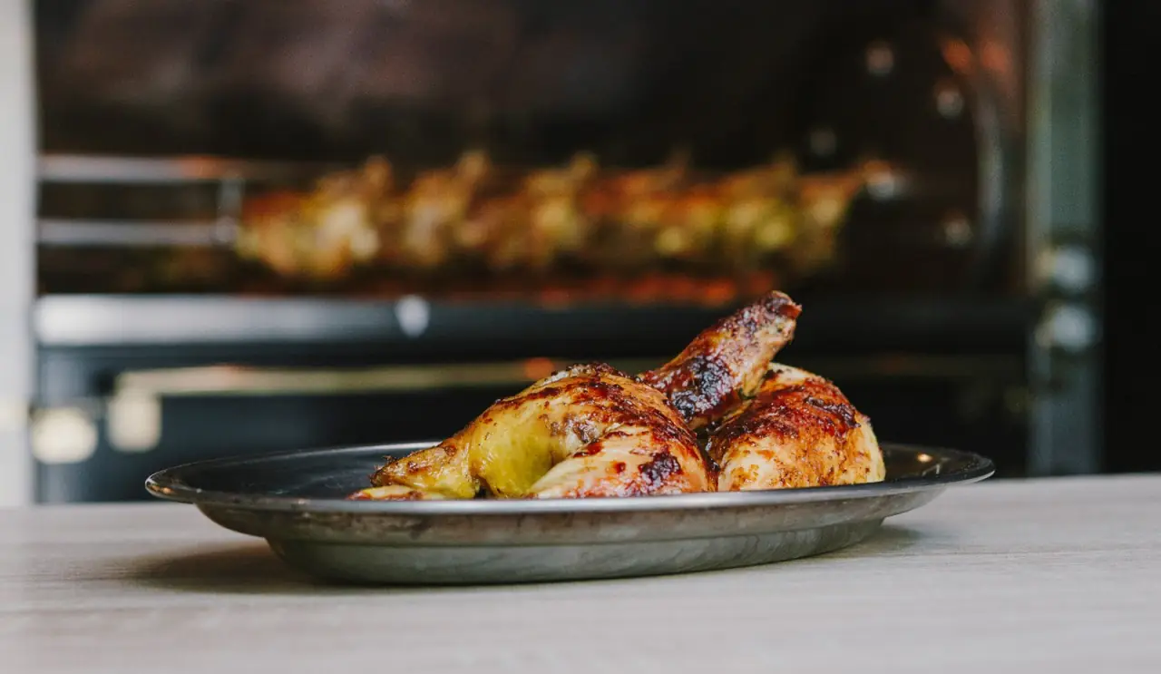Roast chicken halves with charred, glazed skin on a metal plate, with more chicken visible cooking on a rotisserie in the background — A Pluma Santaló