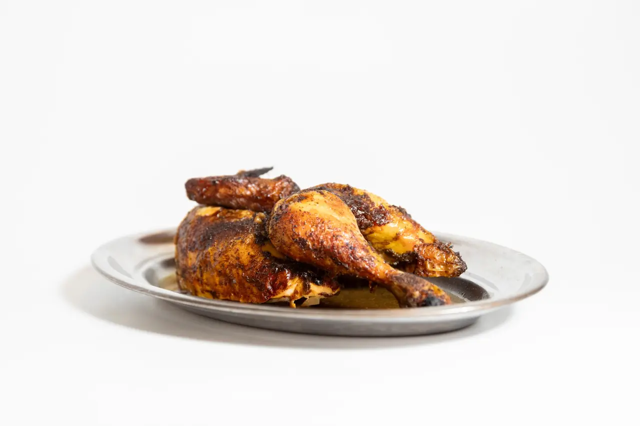 Roast chicken with dark, caramelized skin plated on an oval metal tray against a plain white background — A Pluma Santaló
