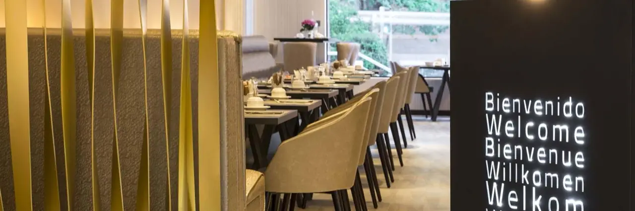 Bright breakfast dining room with a long row of tables set with white place settings and taupe upholstered chairs beside a window