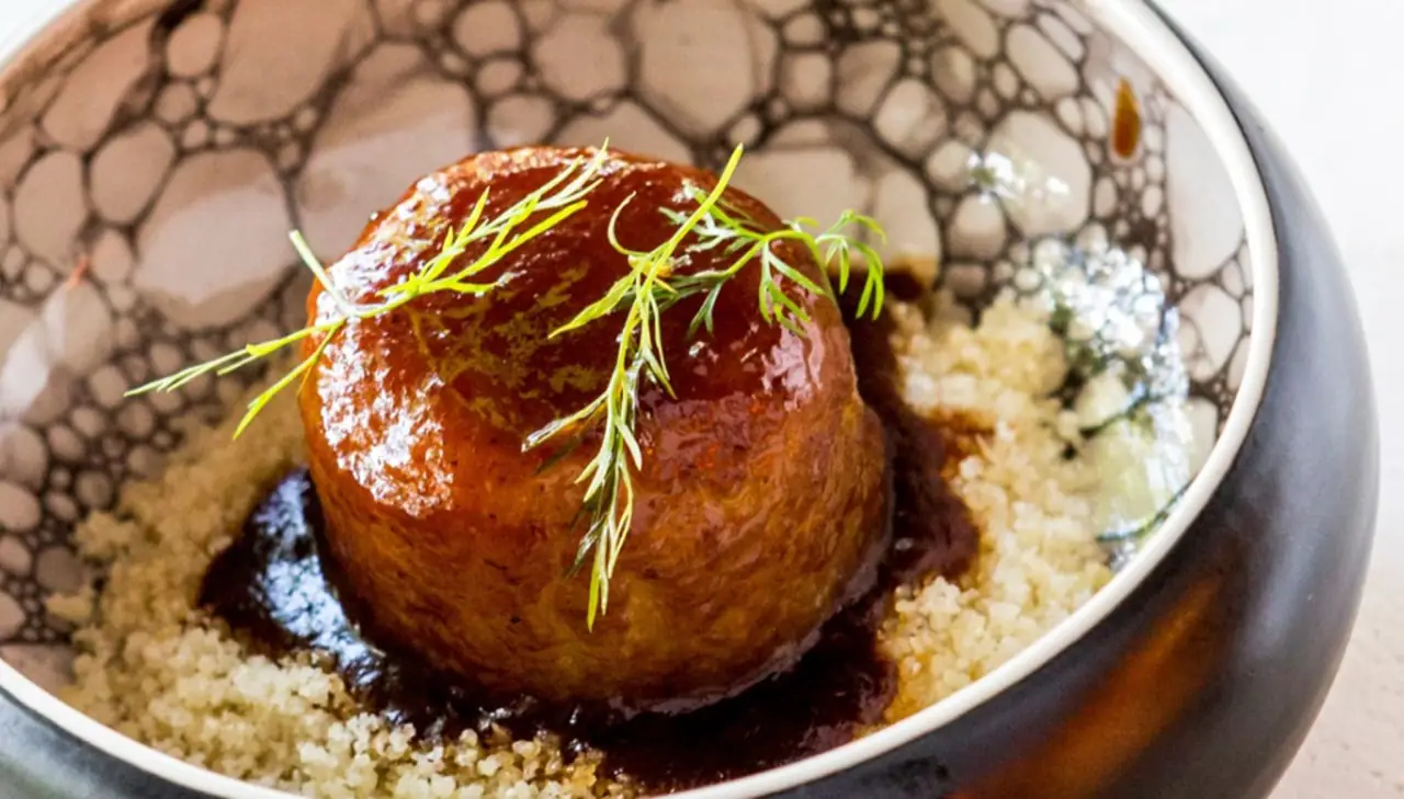 Glazed meatball in dark sauce over couscous, garnished with fresh dill, in a speckled ceramic bowl