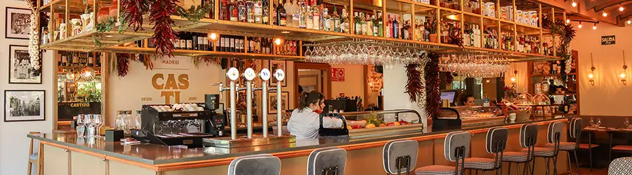 Restaurant bar counter with hanging wine glasses, strings of dried red peppers, a back wall of bottles and bar stools along the front