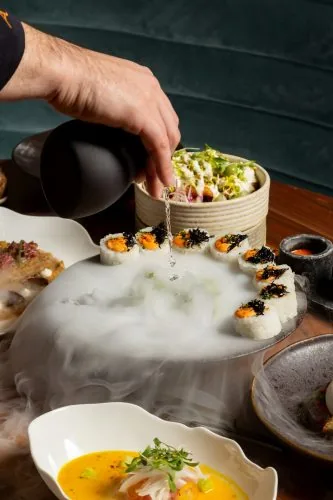 A hand pouring liquid over a raised platter of maki topped with black caviar and orange roe, dry-ice mist spilling across the table, with a tartare bowl and a saffron-coloured soup alongside