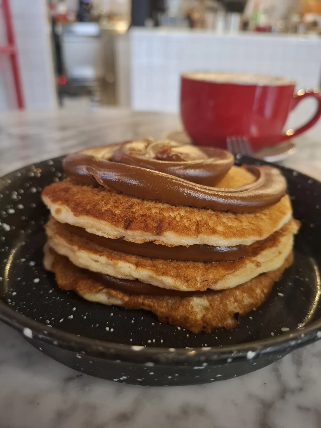 A stack of pancakes drizzled with chocolate spread on a dark speckled plate beside a red coffee cup