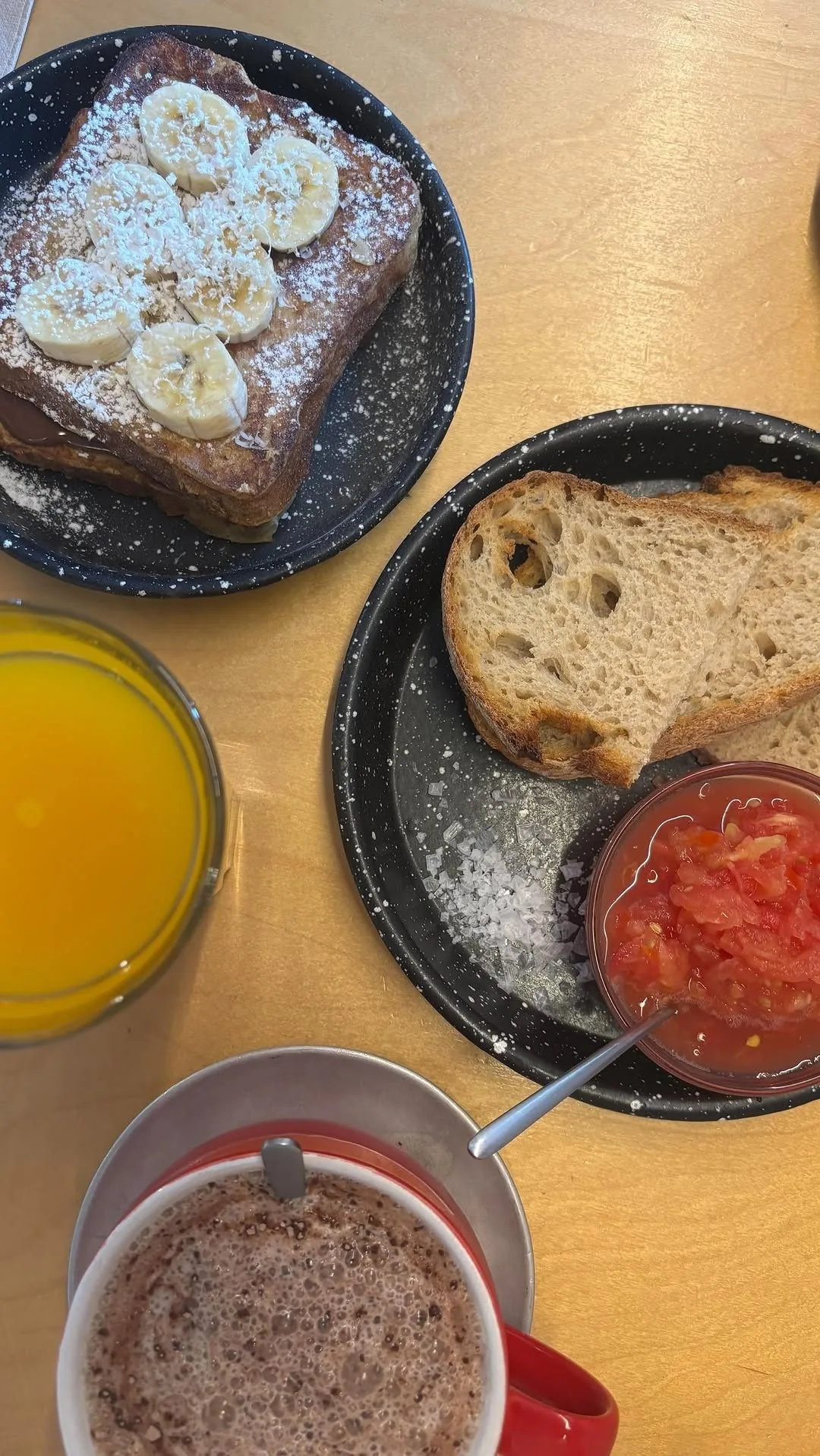 A breakfast table with banana-topped toast, bread, a small pot of tomato, orange juice and a cup of hot drink