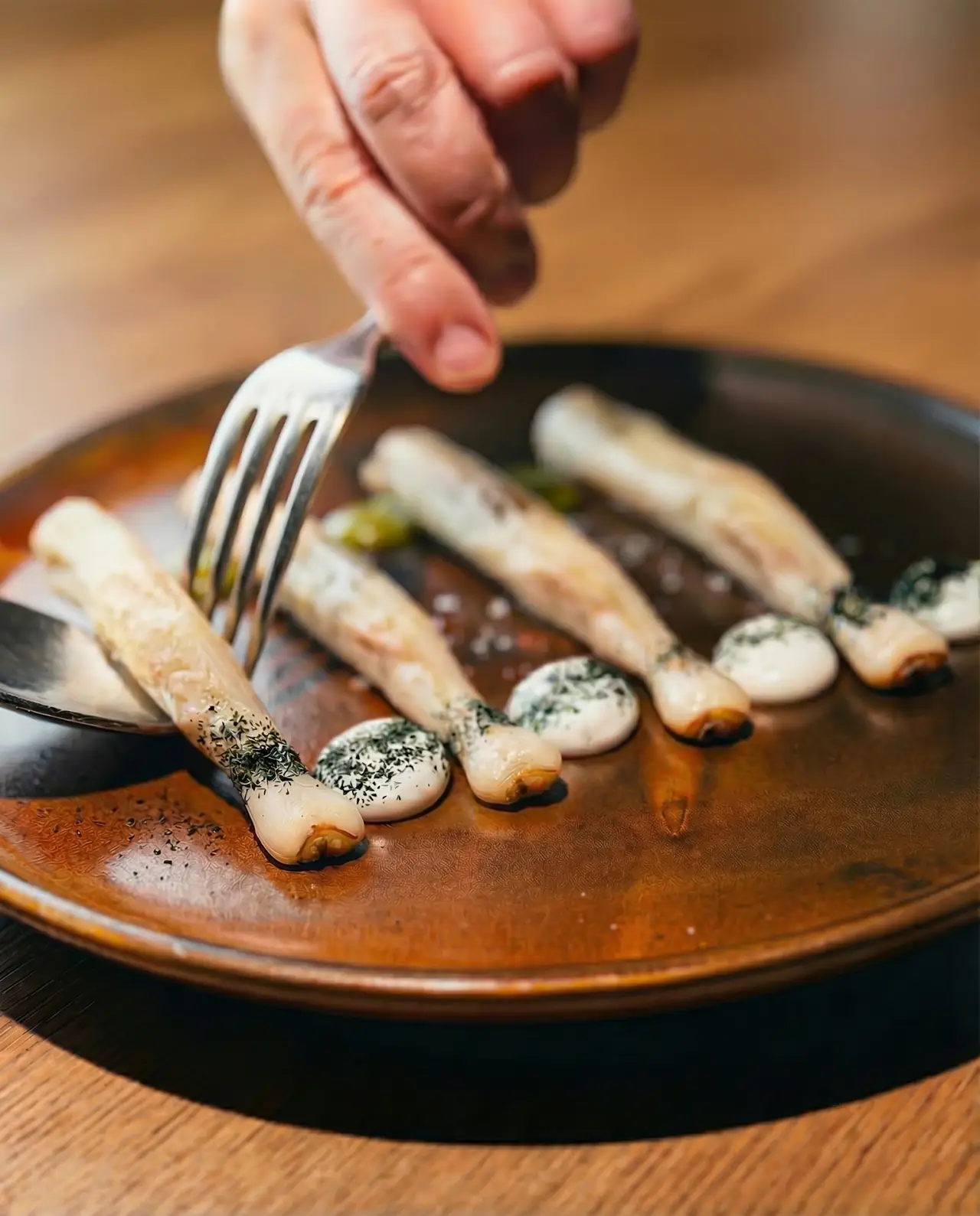 Razor clams plated in a row with herb-flecked sauce dollops and sea salt, a diner reaching in with a fork