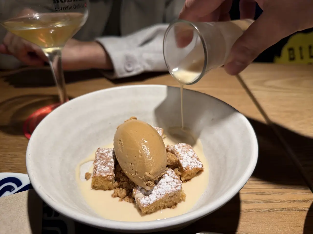 A scoop of caramel ice cream over cake squares in a bowl, with cream being poured tableside