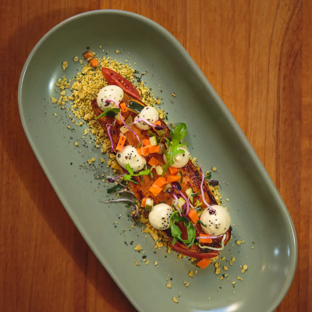 Sliced glazed vegetable dish topped with cashew cream dollops, diced carrot, tomato, sprouts and micro greens on a savory crumb base, served on an oval green plate at Aguaribay