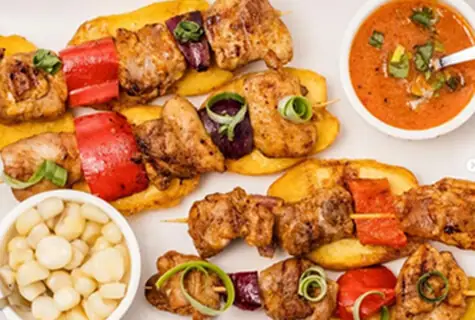 Grilled chicken skewers with red pepper and onion over sliced potatoes, with choclo corn and a dipping sauce