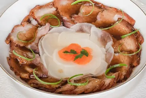 Sliced glazed pork arranged in a ring around radish and carrot garnish with spring onion, on a white plate