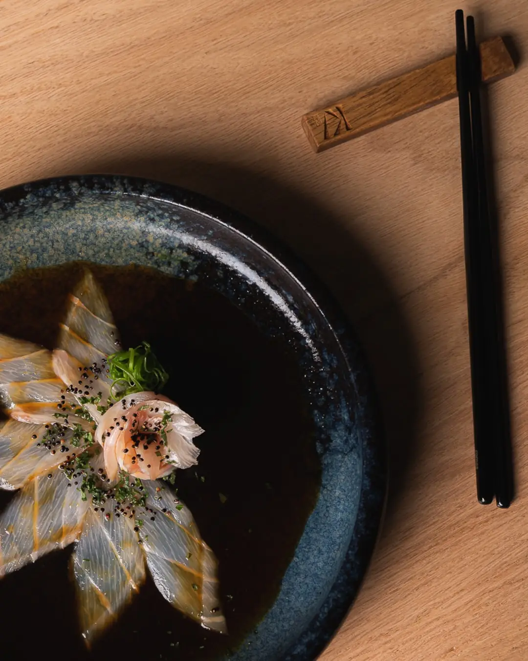 Thinly sliced white fish sashimi fanned on a dark ceramic plate with sauce