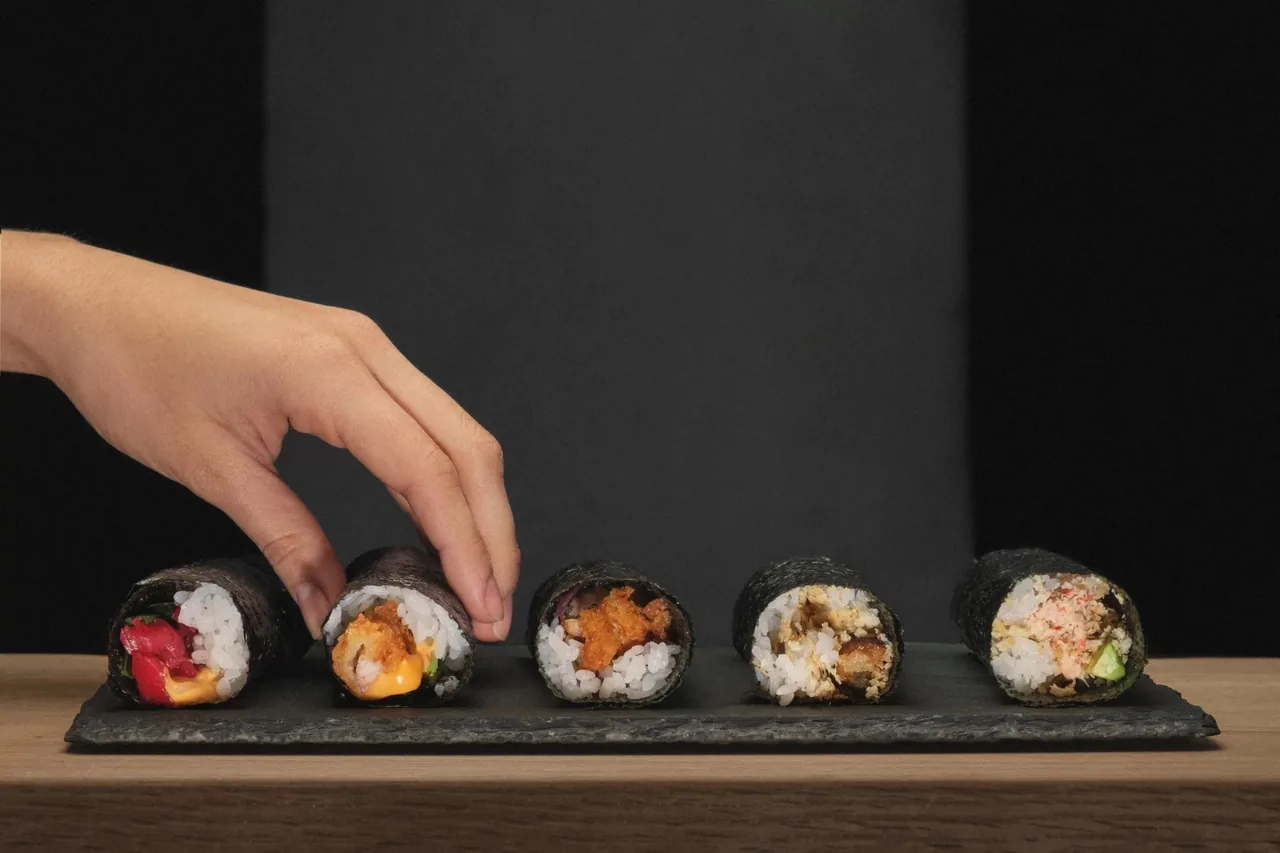 Five hand rolls with different fillings lined up on a slate tray while a hand reaches for one
