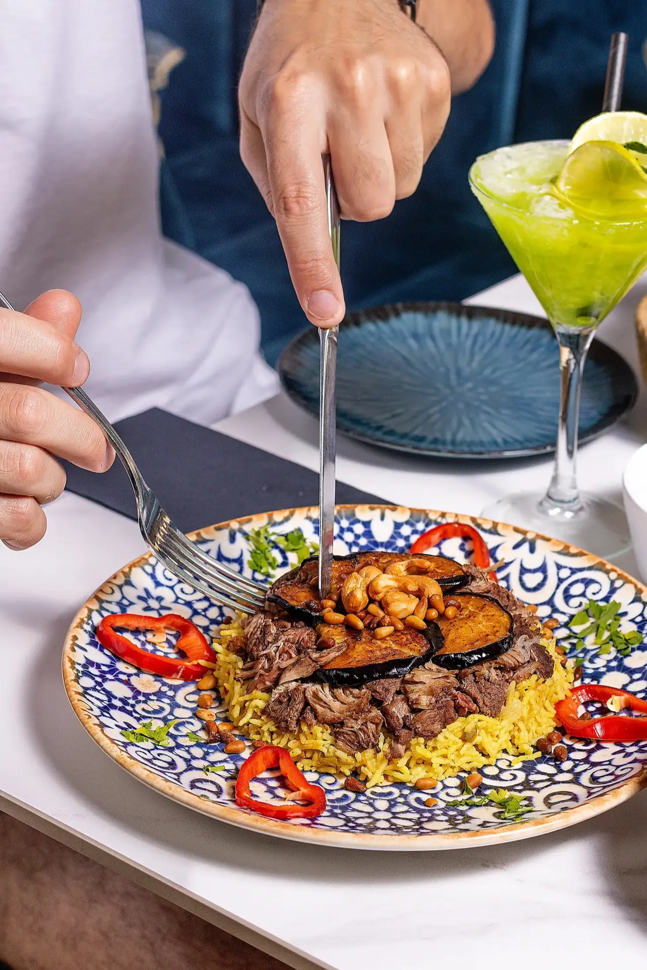 Maqluba-style rice with shredded lamb, fried eggplant and toasted nuts on a blue patterned plate, hands cutting in, with a bright green cocktail.