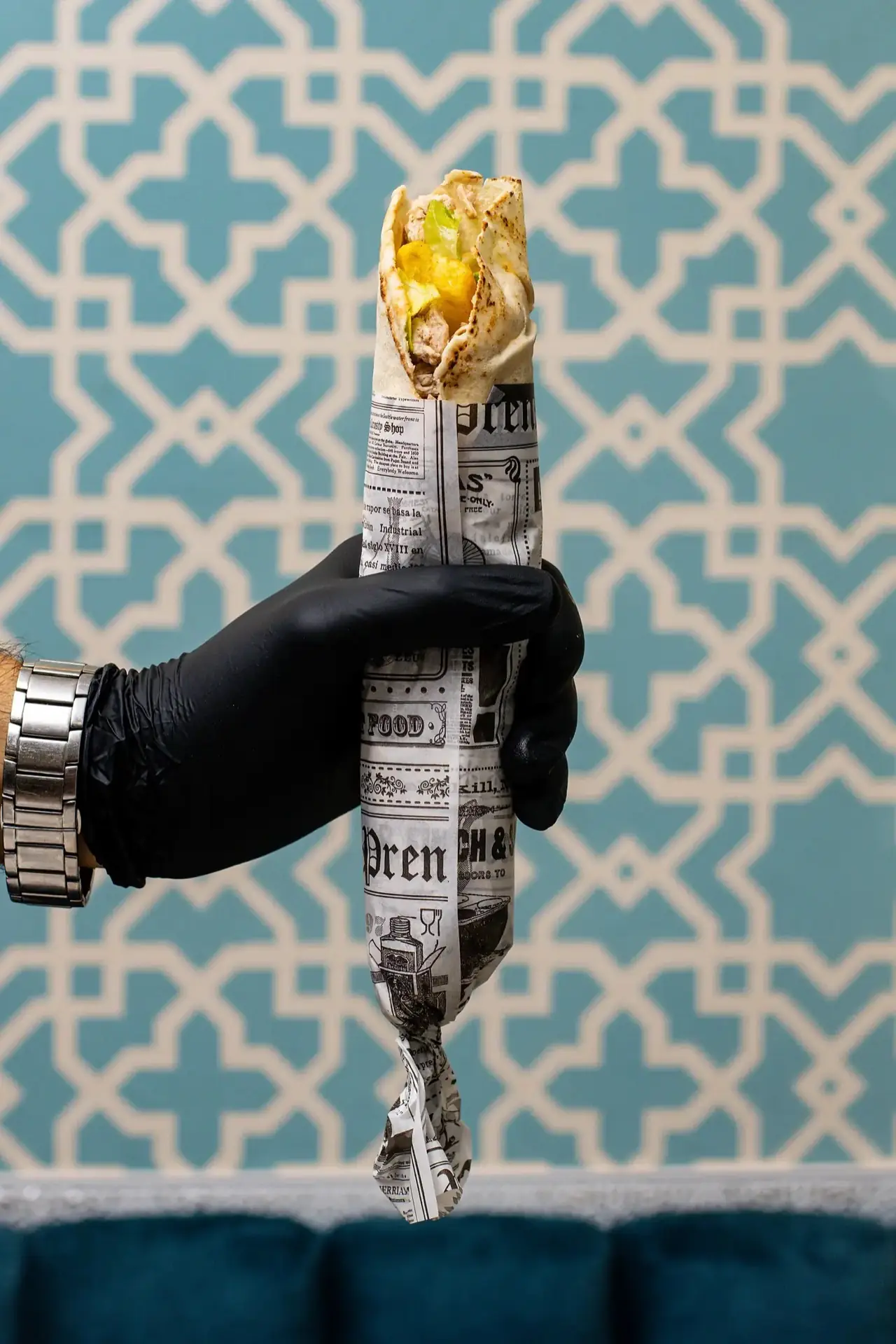 Gloved hand holding a chicken shawarma wrap in newspaper against a blue patterned wall.