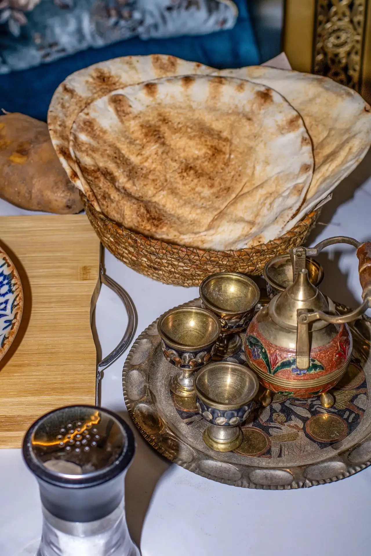 Grilled flatbreads in a woven basket beside a brass tea set with small cups on an engraved tray.