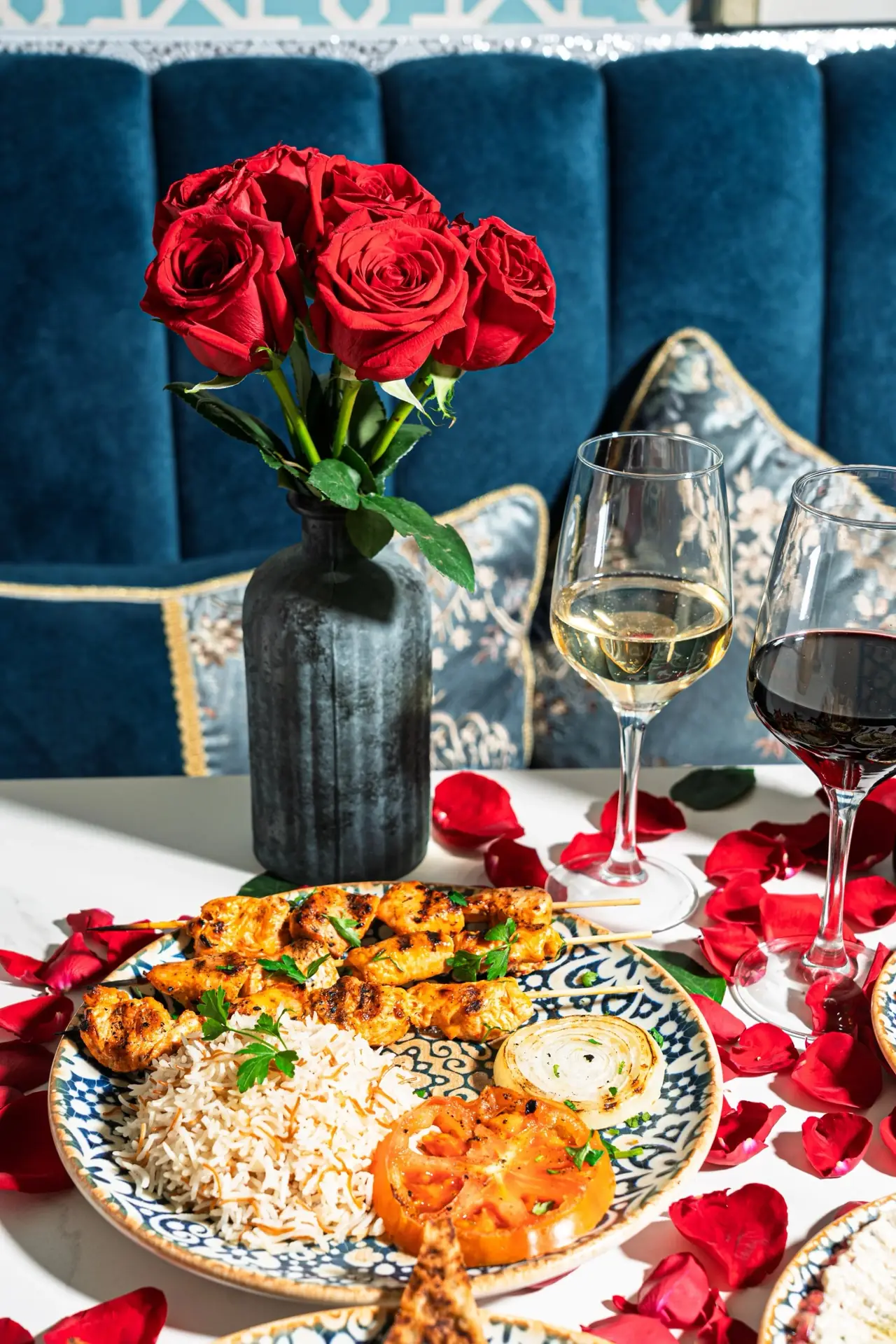 Grilled chicken skewers with rice, grilled tomato and onion on a patterned plate, with glasses of red and white wine and red roses.