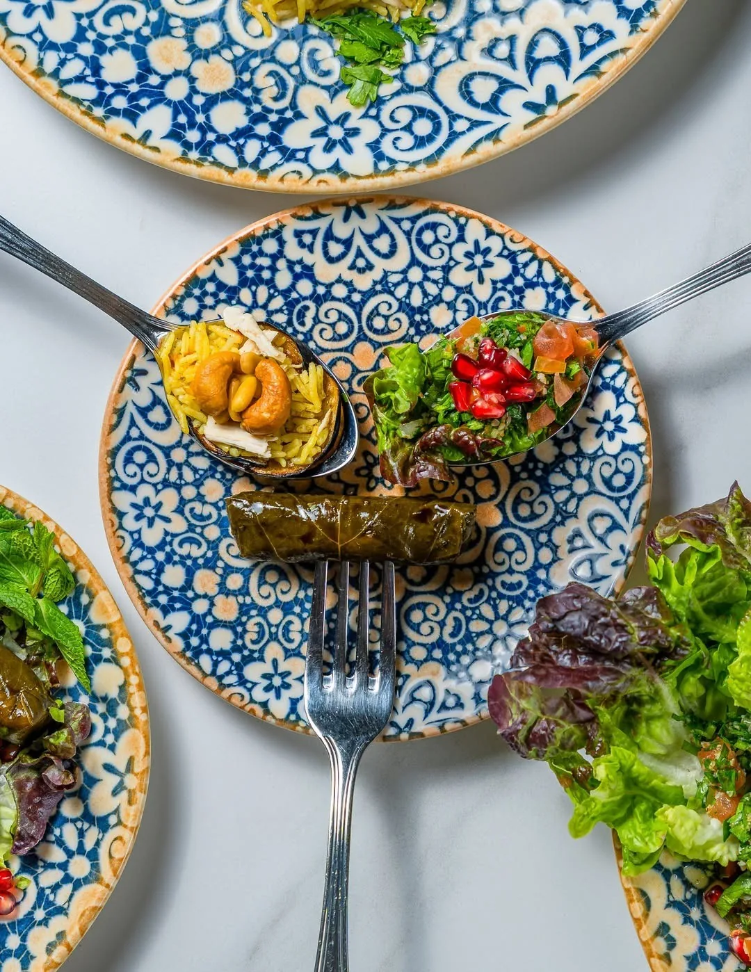 A patterned plate with a spoonful of rice and nuts, a spoonful of chopped salad with pomegranate seeds and a stuffed vine leaf on a fork