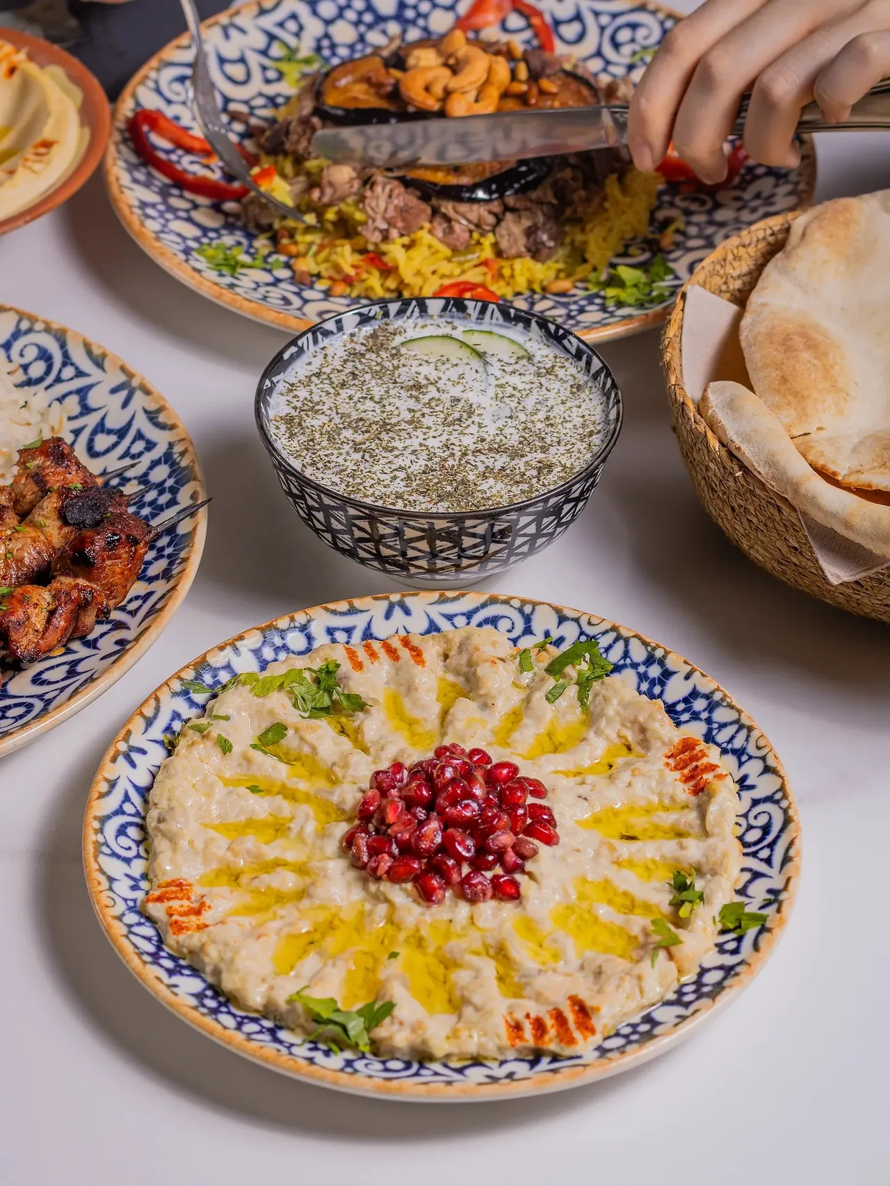A patterned plate of creamy eggplant dip topped with pomegranate seeds, with a yoghurt bowl, grilled meat and a basket of flatbread
