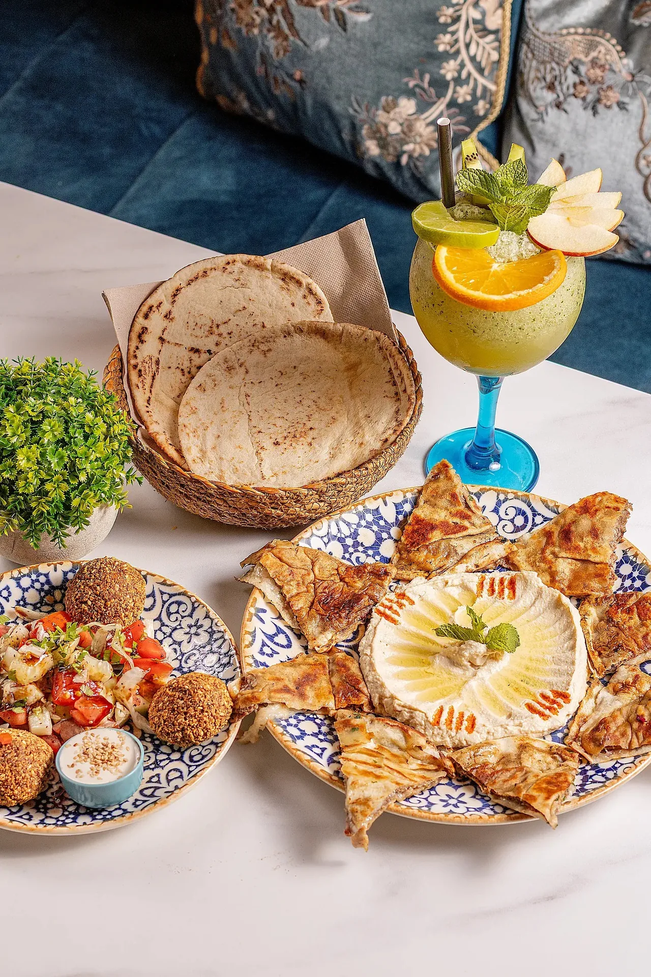 A patterned plate of hummus surrounded by grilled flatbread triangles, with falafel and salad and a basket of pita