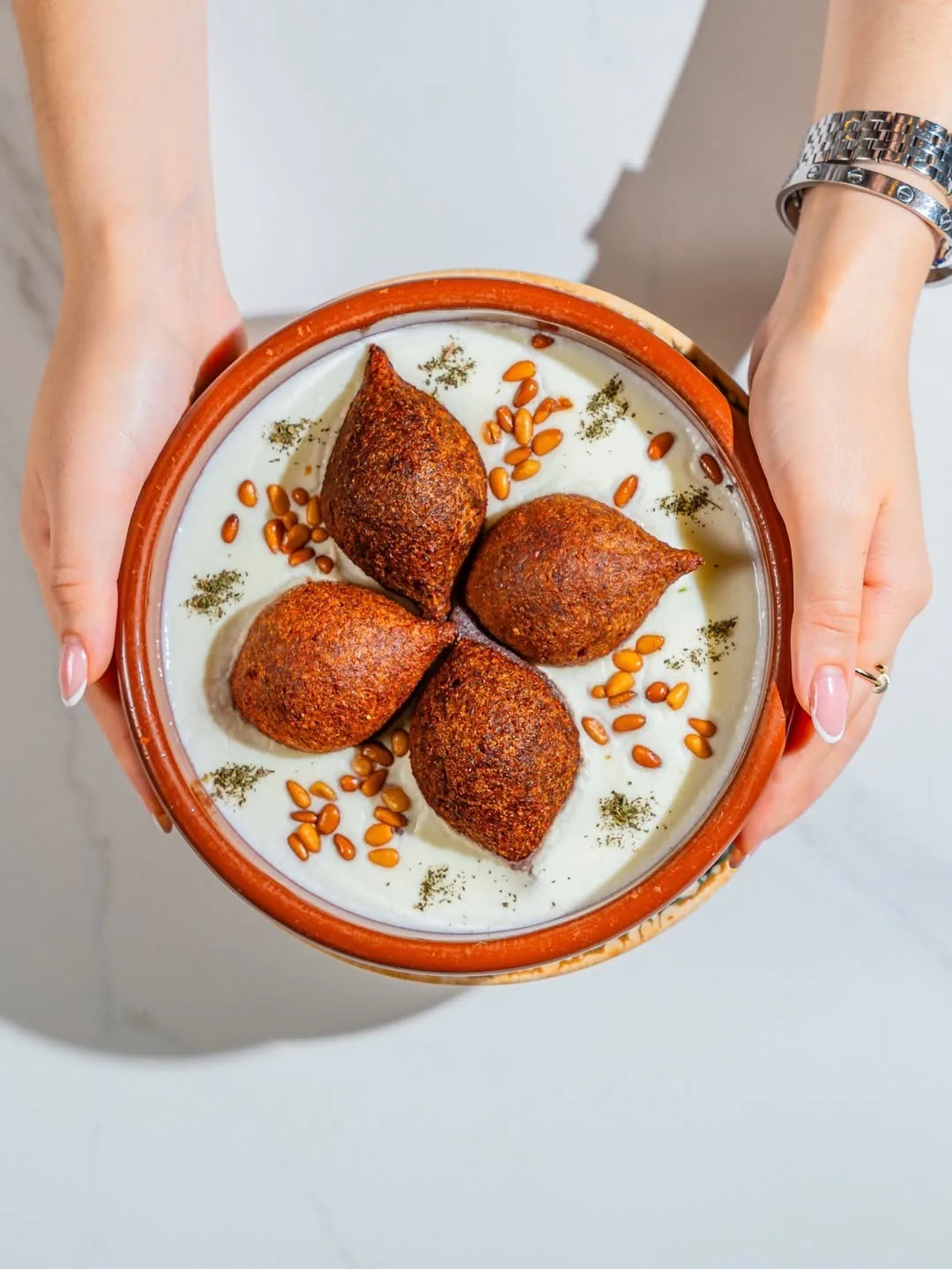 Two hands holding a round dish of fried kibbeh in white sauce