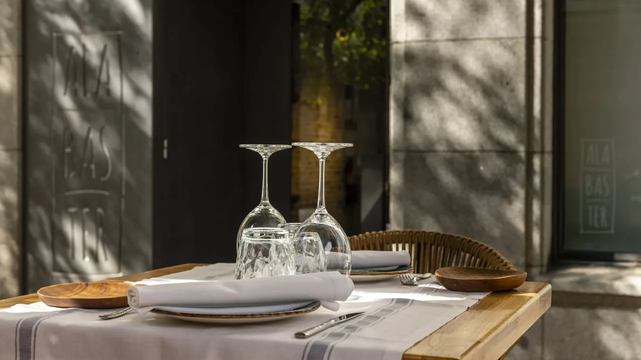 Sunlit terrace table set with upturned wine glasses and wooden plates at Alabaster, the restaurant's name etched on the glass door behind