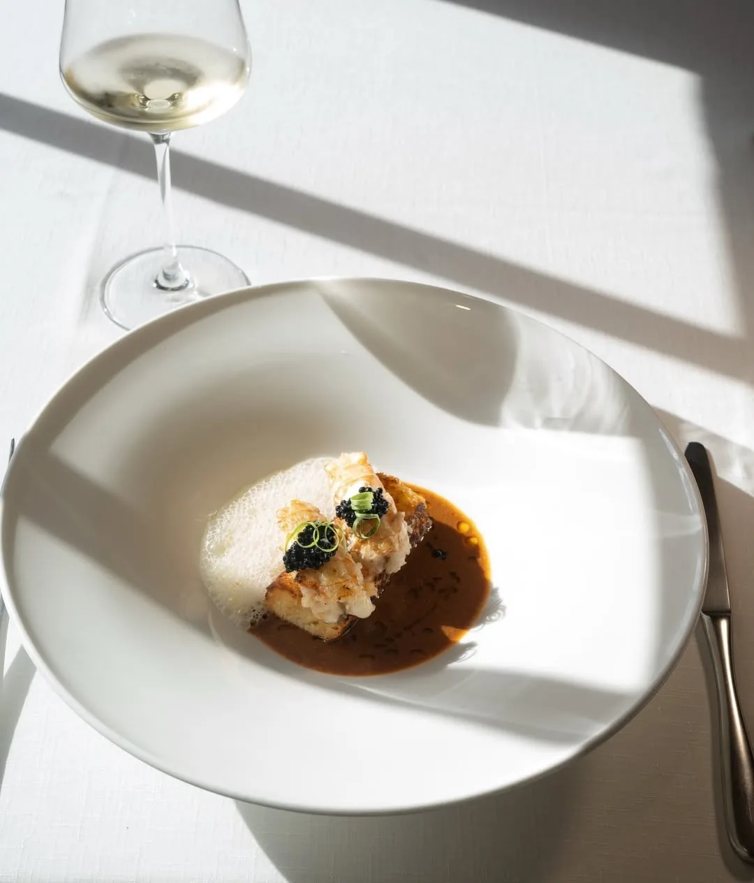 A white bowl of pale fish in a brown sauce with a small green garnish on a white table beside a wine glass