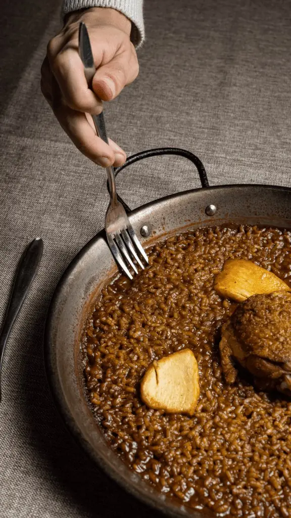 Dark, glossy rice cooked in a metal paella pan topped with meat and roasted garlic cloves, a diner's hand holding a fork over it