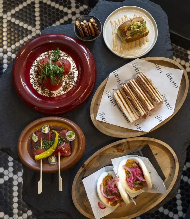 Overhead spread of small plates including bao buns, a striped club sandwich on branded paper, a red-plate tartare and tuna crudo on a dark tiled surface