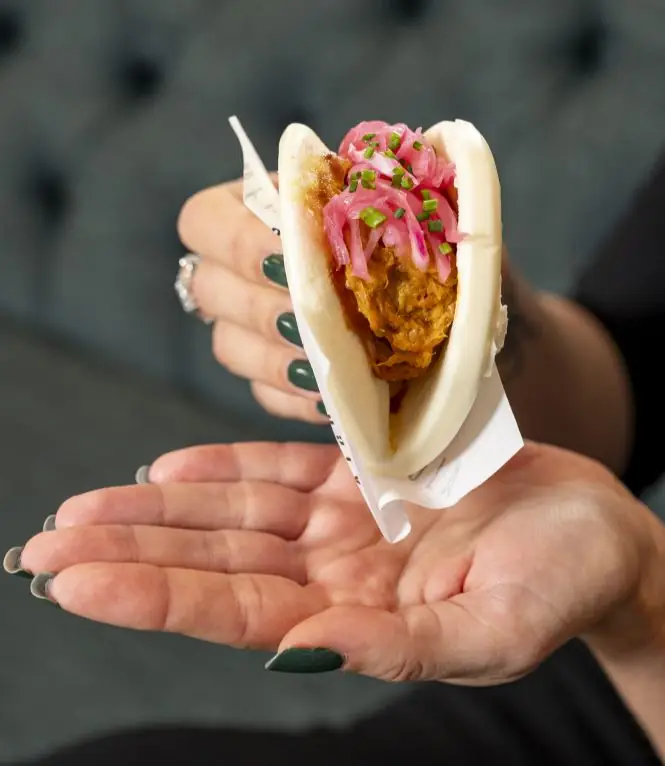A hand holding a bao bun filled with fried chicken and topped with pickled red onion and chives