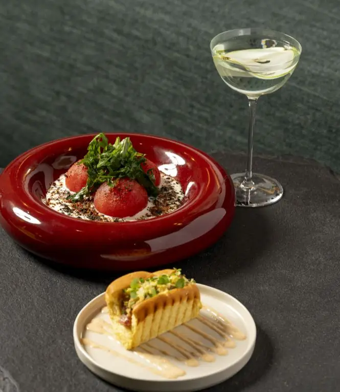 A red bowl of tomato and cream dish and a slice of tart on a plate beside a coupe cocktail on a dark green background