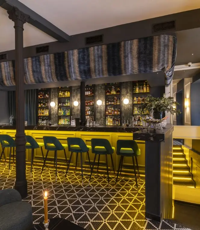Dimly lit bar with green velvet stools along a yellow-fronted counter, a back bar of backlit bottles and a patterned mosaic floor