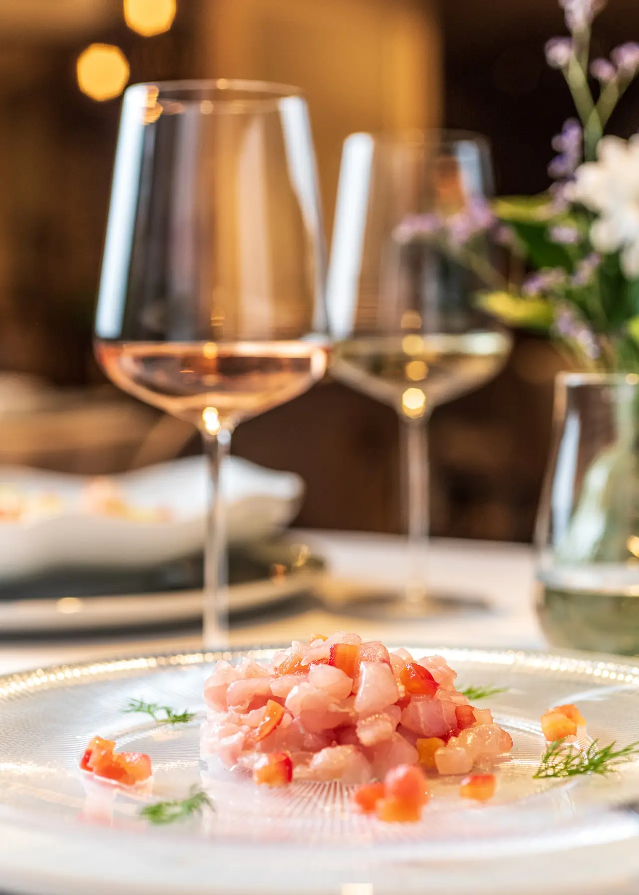 Diced fish tartare with tomato and fennel on a glass plate, wine glasses in the background