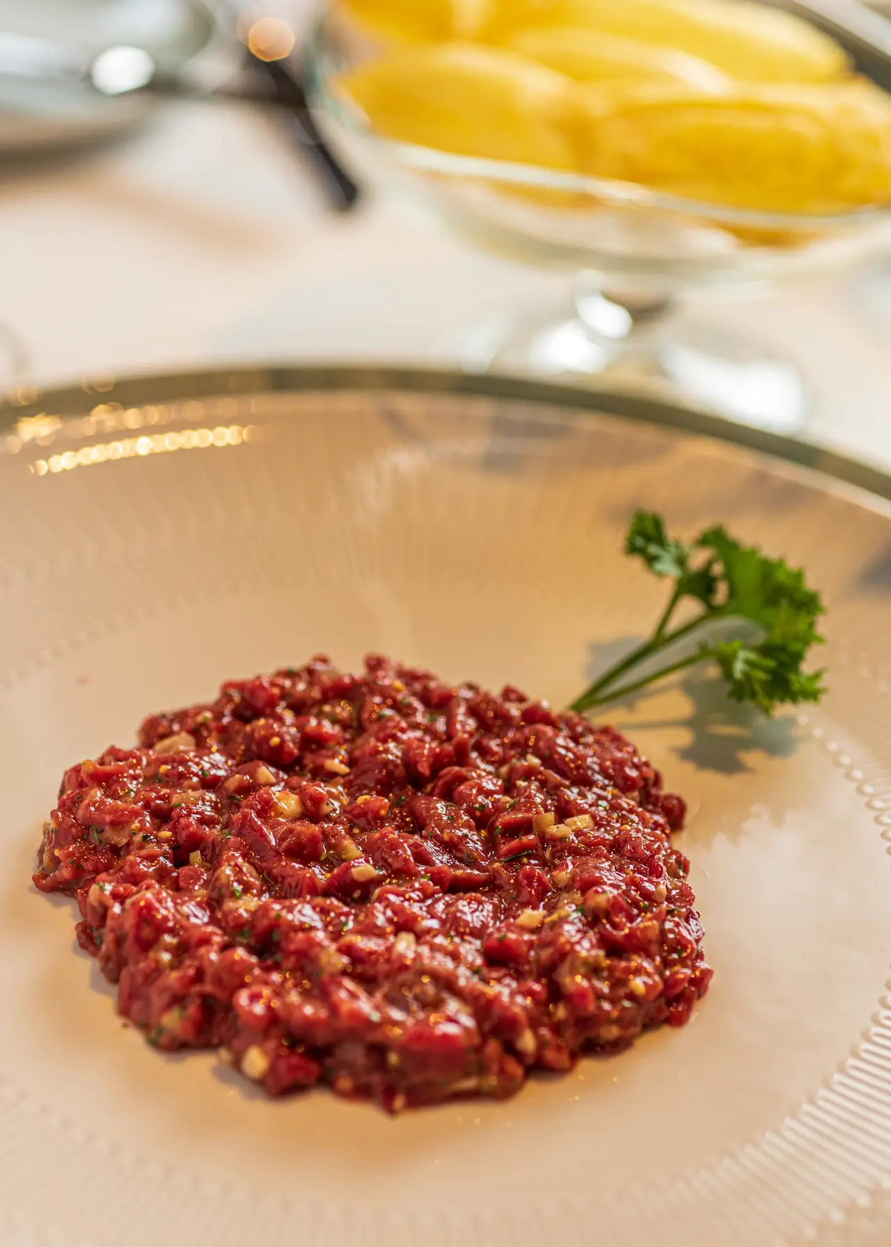 Beef tartare with a parsley garnish on a glass plate, bowl of potato chips in the background