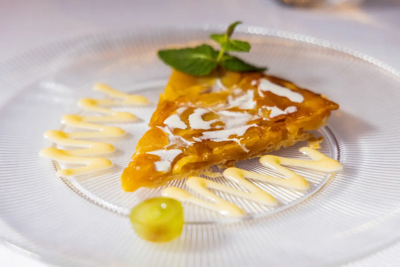 Slice of apple tart with a cream drizzle, mint sprig and a grape on a glass plate