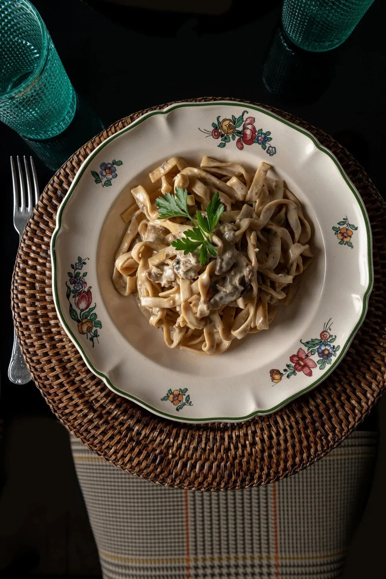 A pasta dish with herbs in a floral-rimmed bowl on a wicker mat