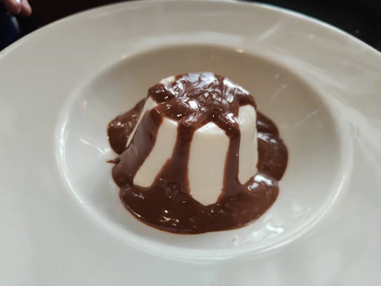 A panna cotta drizzled with chocolate sauce on a white plate.