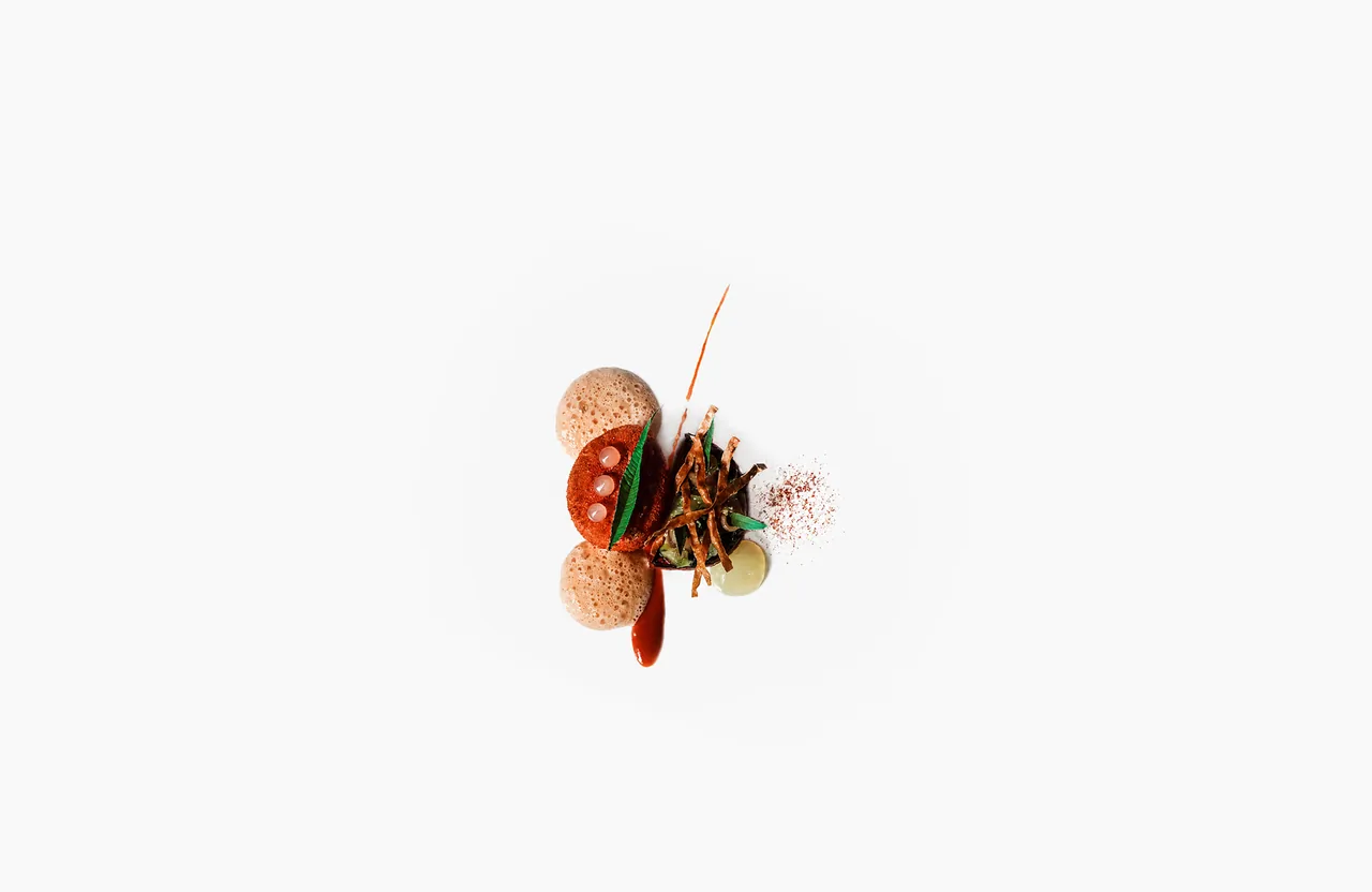 A small fine-dining plate shot from directly above on a plain white surface: a crimson crumb-crusted disc dotted with three pale pink pearls and pierced by a gr