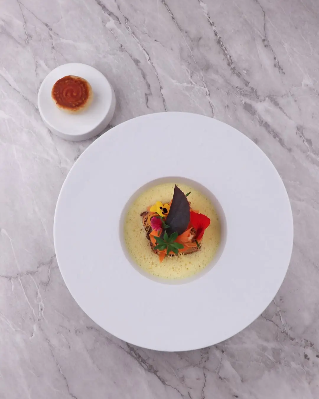 Round plated dish with a saffron-colored foam sauce, herbs and edible flowers beside a small side bowl